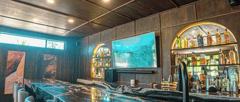 A bar area with a marble countertop, bar stools, a television, and shelves of liquor bottles.