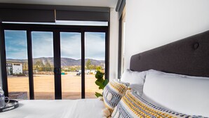 A modern hotel room with a large bed, a view of a parking lot and mountains, and a window with blinds. - Quinta Nicolás Valle de Guadalupe (Valle de Guadalupe)