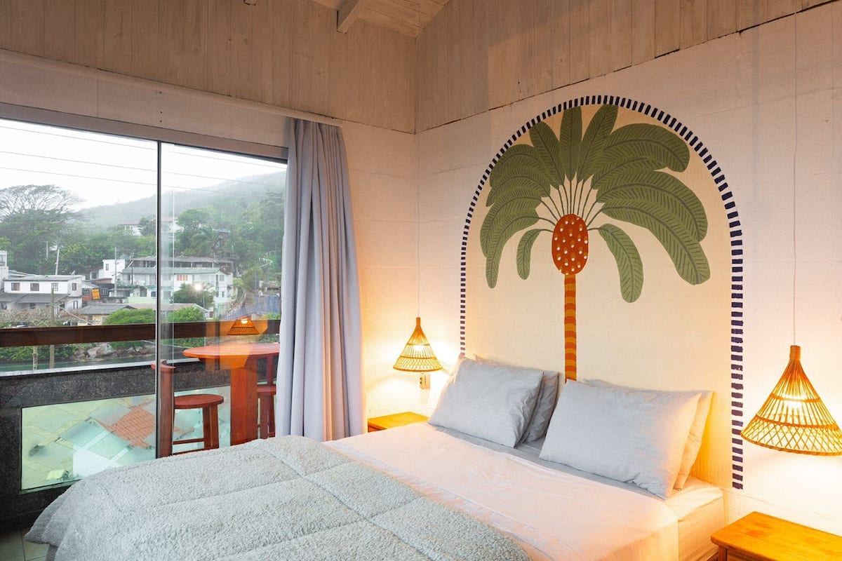 Un dormitorio con una cama grande, dos lámparas de noche, vistas a un pueblo por la ventana y un mural de una palmera.