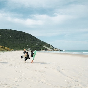 A sandy beach with footprints leading towards the ocean, a green hill in the background, and a cloudy sky. - Sea Wolf Surf Club - Hostel (Florianópolis)