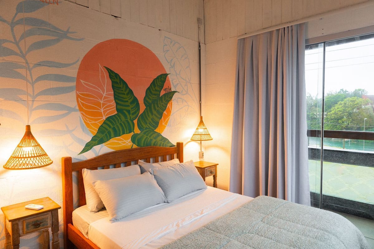 Un dormitorio con una cama, dos lámparas de noche, un ventanal con cortinas y un mural en la pared.