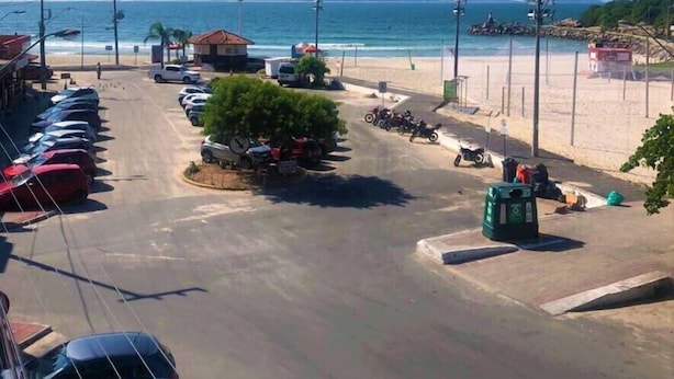 Un estacionamiento con coches y motocicletas, una playa con sombrillas y una vista despejada del mar y las montañas al fondo.