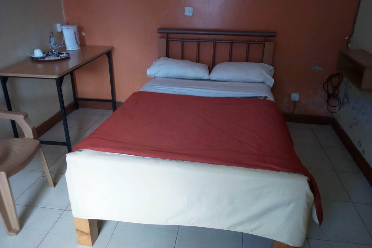 A single-bed room with a red blanket, a wooden desk, a chair, and a wall socket.