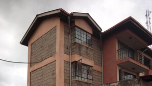Un edificio de dos pisos con techo plano, un balcón en el segundo piso y una estructura pequeña adosada con un ventanal.