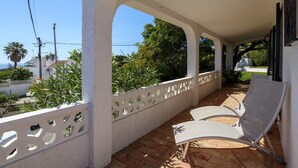 Ein überdachter Außenbereich mit weißen Wänden und einem weißen Gitterzaun, ausgestattet mit zwei weißen Lounge-Stühlen und Blick auf Grünflächen und Gebäude.