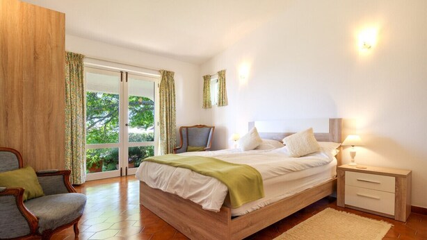 Ein Schlafzimmer mit einem hölzernen Bett, einem Nachttisch, einem Sessel und Blick auf Grün hinter einer Schiebetür.
