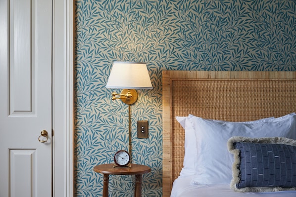 A bedroom with a patterned wallpaper, a bedside lamp, a clock, and a bed with pillows.