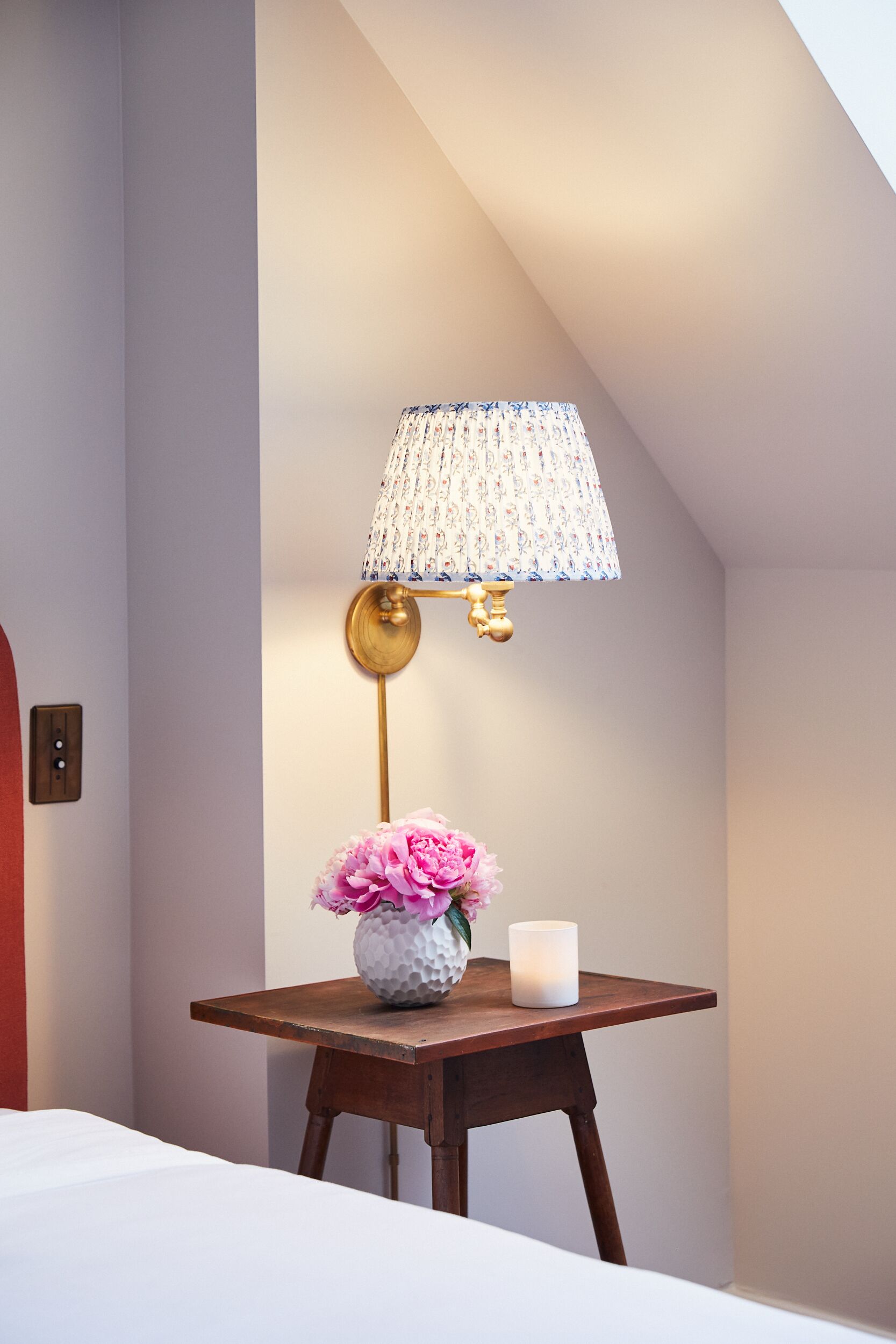 A corner of a room with a wooden table, a vase with pink flowers, a white candle, and a lamp with a patterned lampshade.