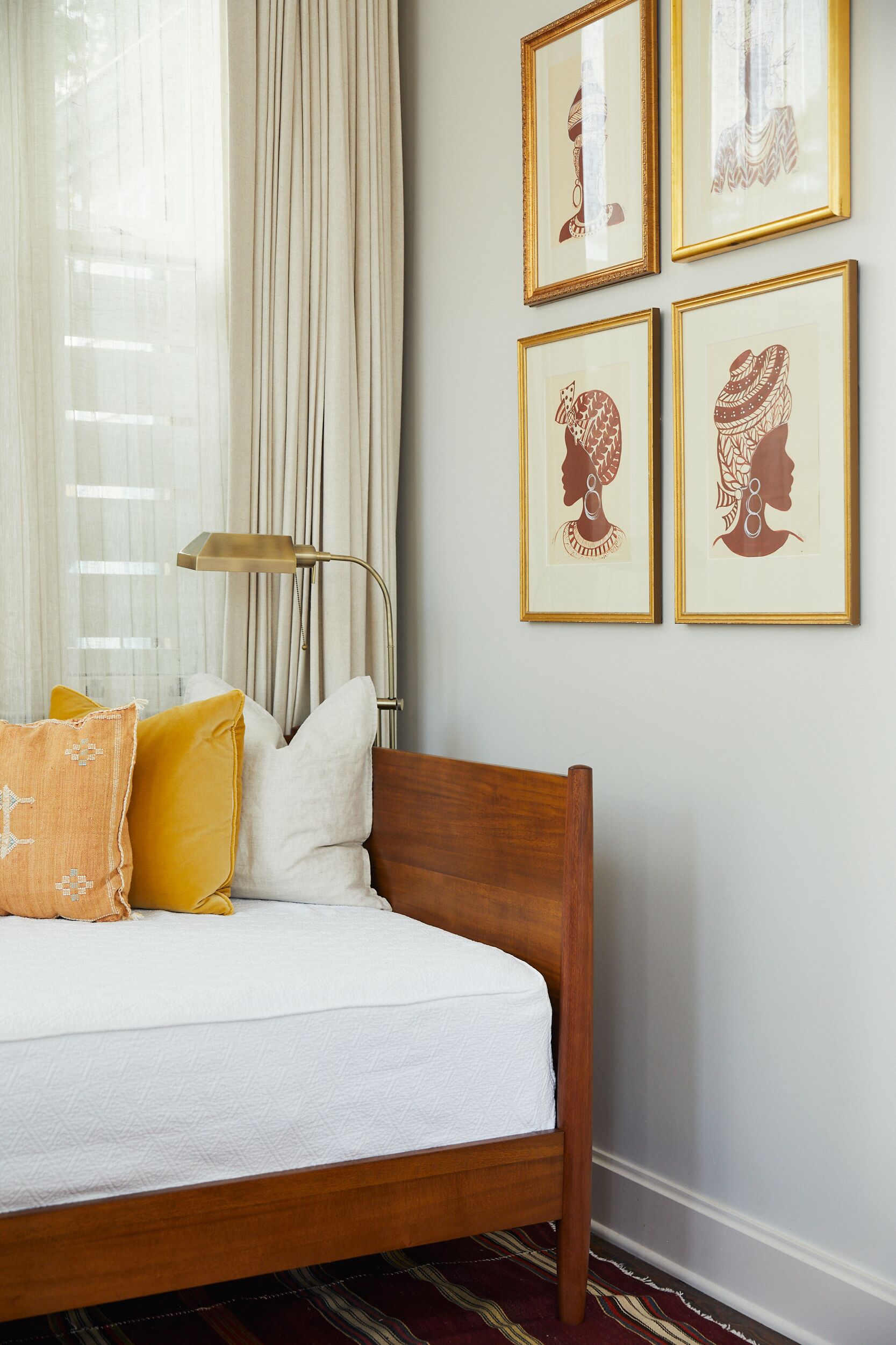 Een net opgemaakt bed met houten frame, witte beddengoed en twee decoratieve kussens. Naast het bed staat een vloerlamp, en er is een raam met dunne gordijnen in de achtergrond zichtbaar.