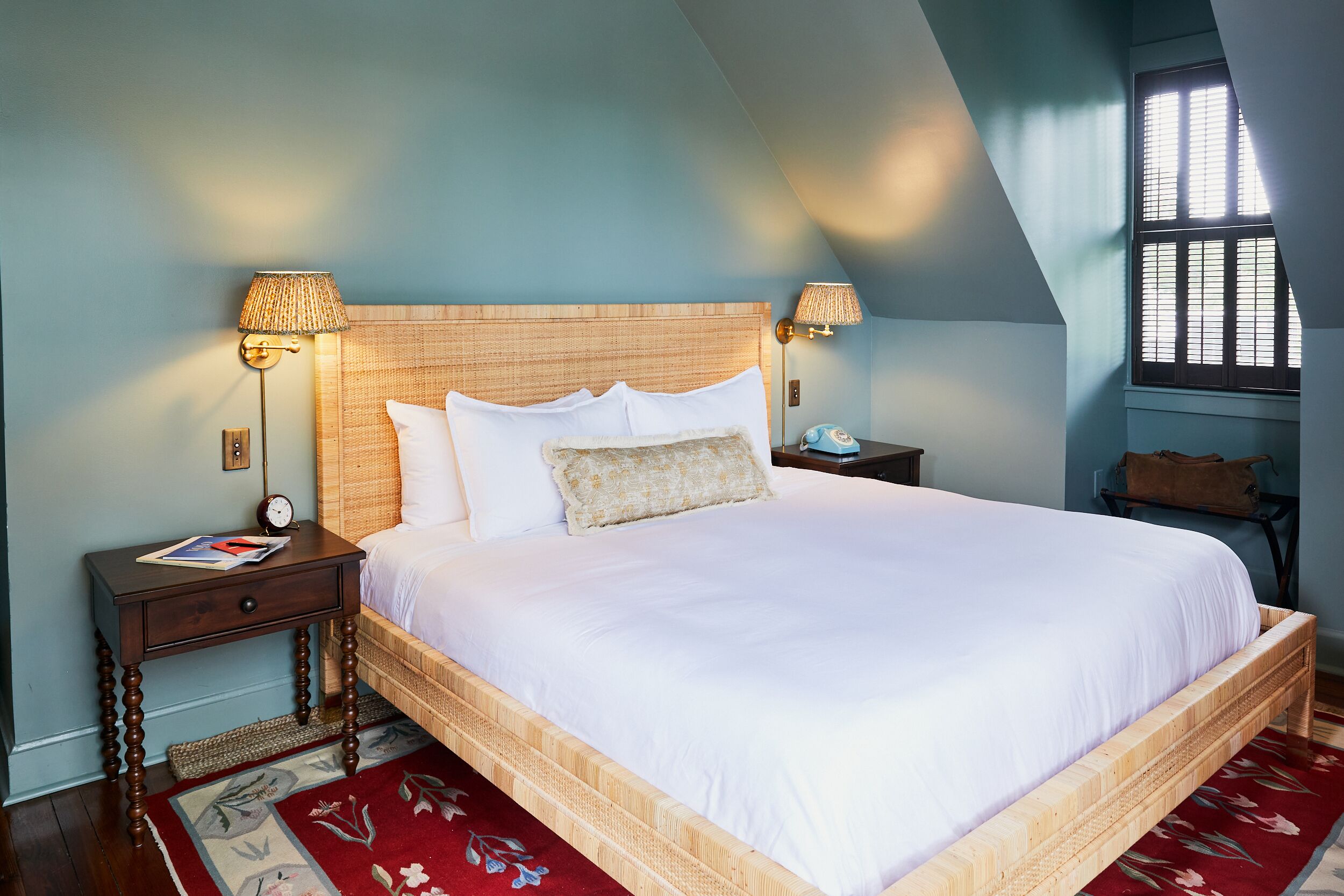 Een opgemaakt bed met een houten hoofdbord, twee nachtkastjes met lampen, een rood bloemenkleed en een raam met zonwering.