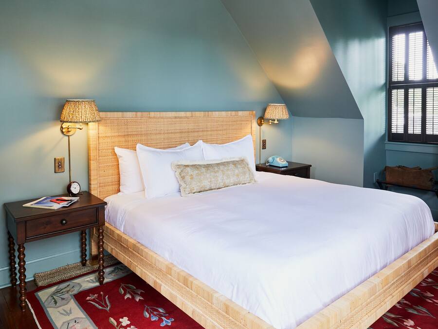 A neatly made bed with a wooden headboard, two bedside tables with lamps, a red floral rug, and a window with blinds.