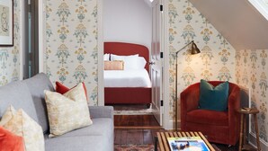 Une pièce chaleureuse avec un canapé gris, un fauteuil rouge, une table basse en bois et un tapis à motifs. On aperçoit un lit avec une literie blanche à travers une porte ouverte.