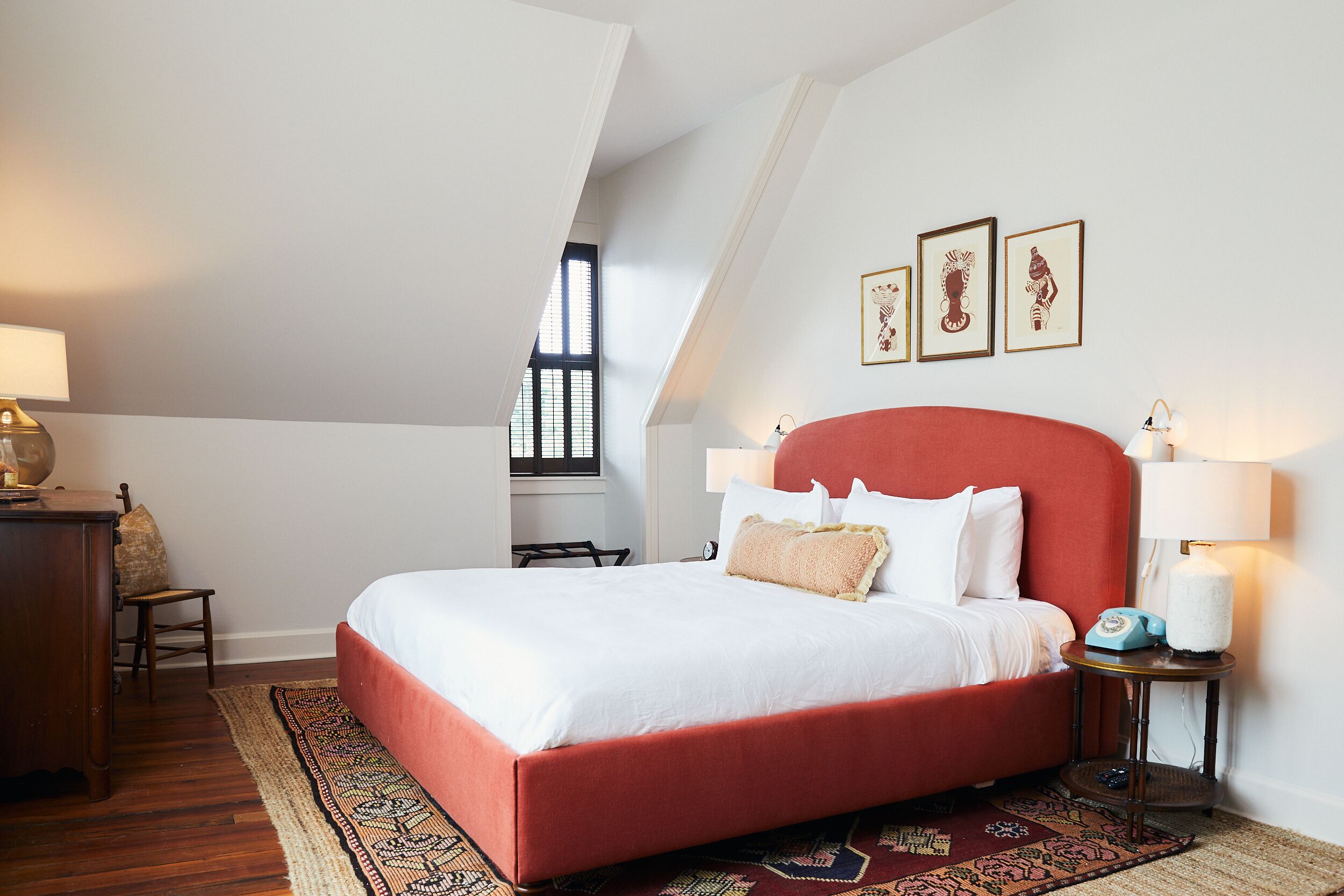 Une chambre à coucher avec un lit rouge, du linge de lit blanc, une commode en bois, une chaise et une fenêtre avec des stores.