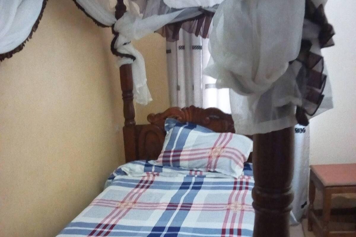 A single bed with a plaid bedspread, a wooden headboard, and a brown bench at the foot of the bed.