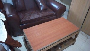 Un canapé en cuir marron, accompagné d’une table basse en bois, se trouve dans une pièce dont le sol est recouvert de carreaux blancs.
