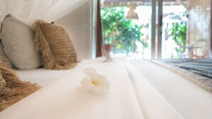 A bed with white linens and a single white flower, a textured brown pillow, and a view of greenery outside. - The Chillhouse Canggu (Canggu)