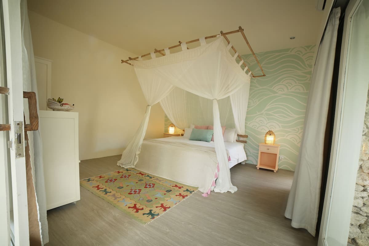 A bedroom with a four-poster bed, a bedside table with a lamp, a patterned rug, and a stone wall.