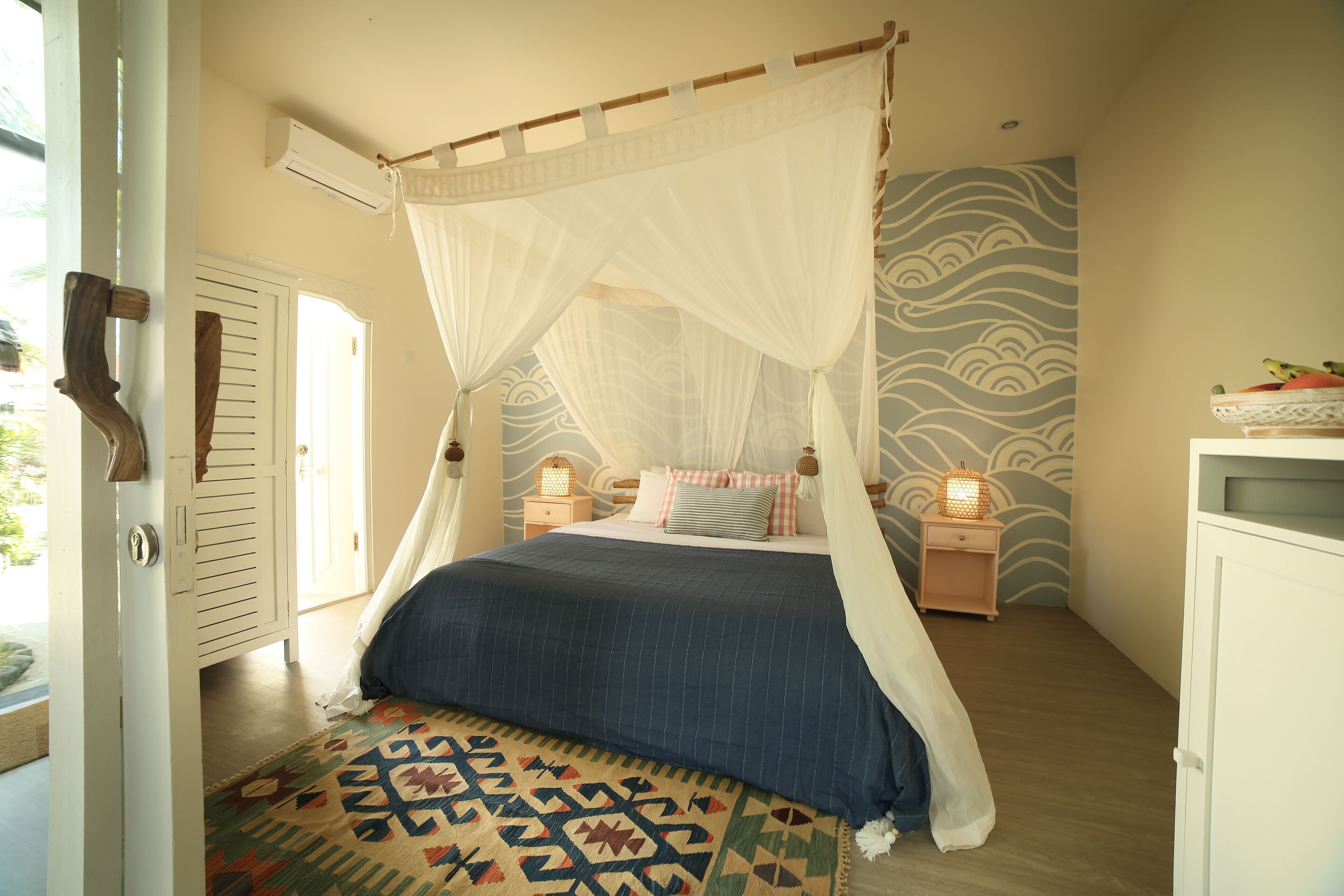 A bedroom with a four-poster bed, a patterned rug, a nightstand with a lamp, and a wall with a wave pattern.
