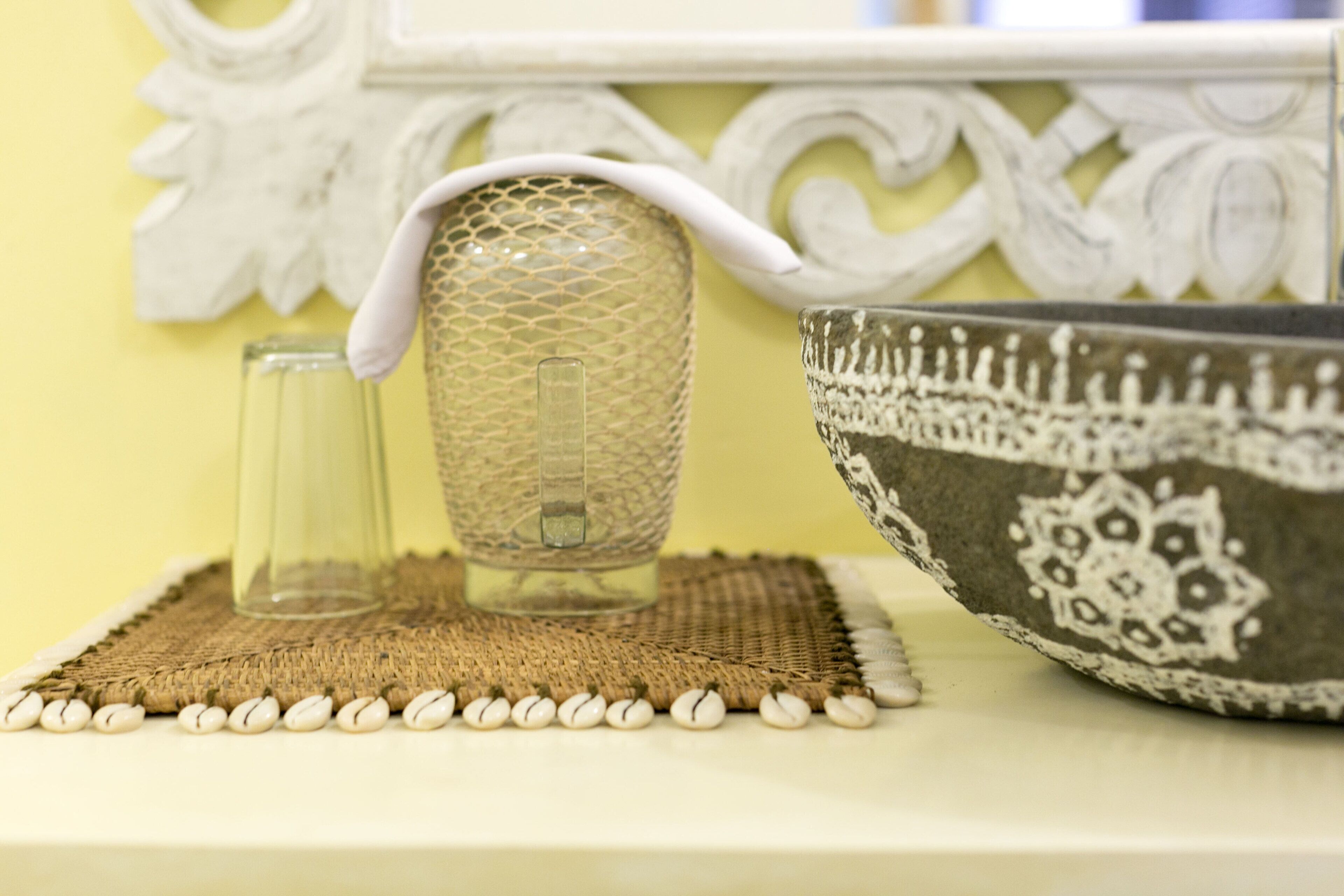 Une carafe en verre décorative, un verre transparent et un bol à motifs, le tout posé sur un set de table en fibres tressées.