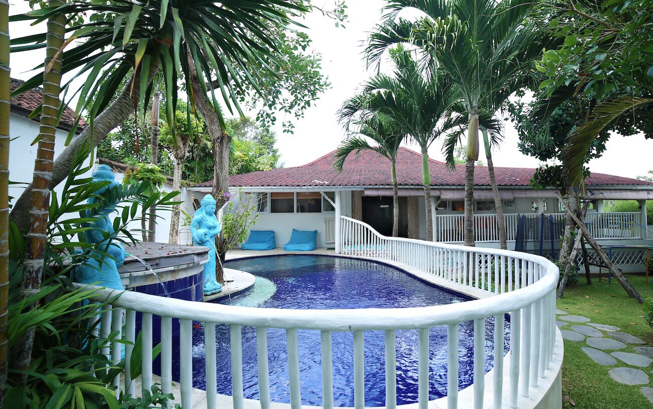 A swimming pool with a white railing, surrounded by palm trees and a house with a red roof.
