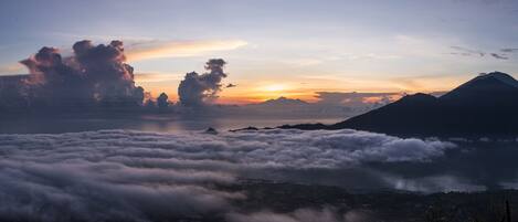 A mountainous landscape with clouds below the peak and a sunset in the background.