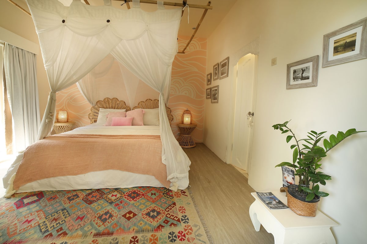 A bedroom with a four-poster bed, a patterned rug, a bedside table with a plant, and framed pictures on the wall.