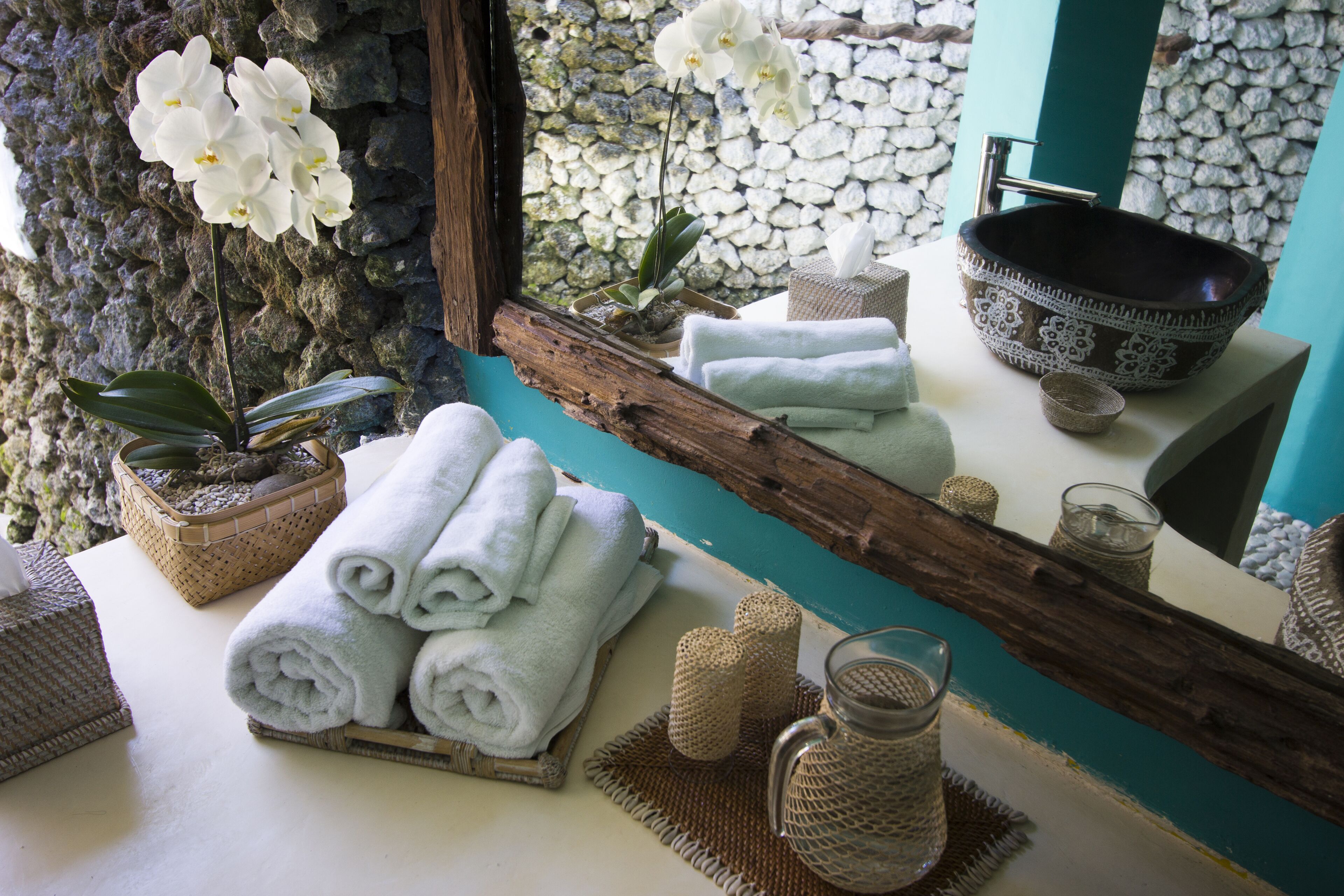 Une salle de bain avec un mur en pierre, un miroir en bois, un lavabo agrémenté d’un récipient décoratif, des serviettes, une orchidée en pot et un panier tressé.