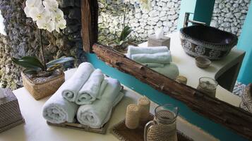 Un baño con pared de piedra, un espejo de madera, un lavabo con un cuenco decorativo, toallas, una orquídea en maceta y una cesta tejida.