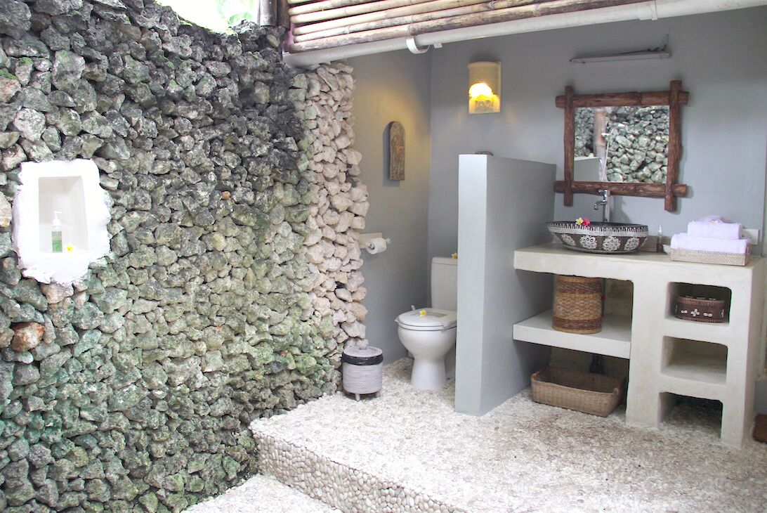 Une salle de bain avec un mur en pierre, un lavabo blanc agrémenté d’un récipient décoratif, un miroir et des toilettes.