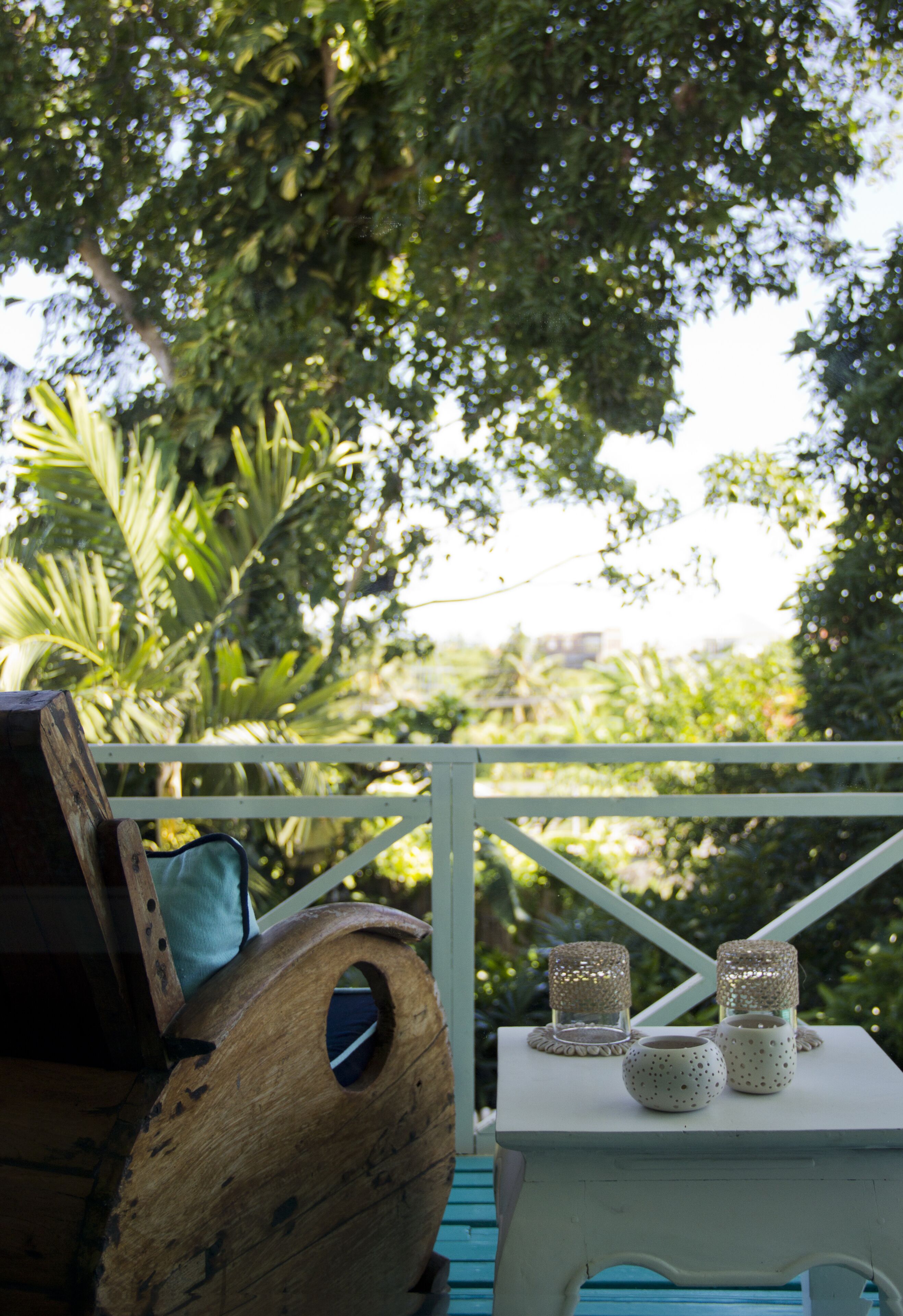 A wooden bench with a blue cushion, a white table with three candle holders, and a view of lush greenery.