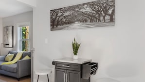 Ein modernes Wohnzimmer mit einem grauen Sofa, einer kleinen Küchenzeile mit einer Pflanze und einem großen Schwarz-Weiß-Baumfoto an der Wand.