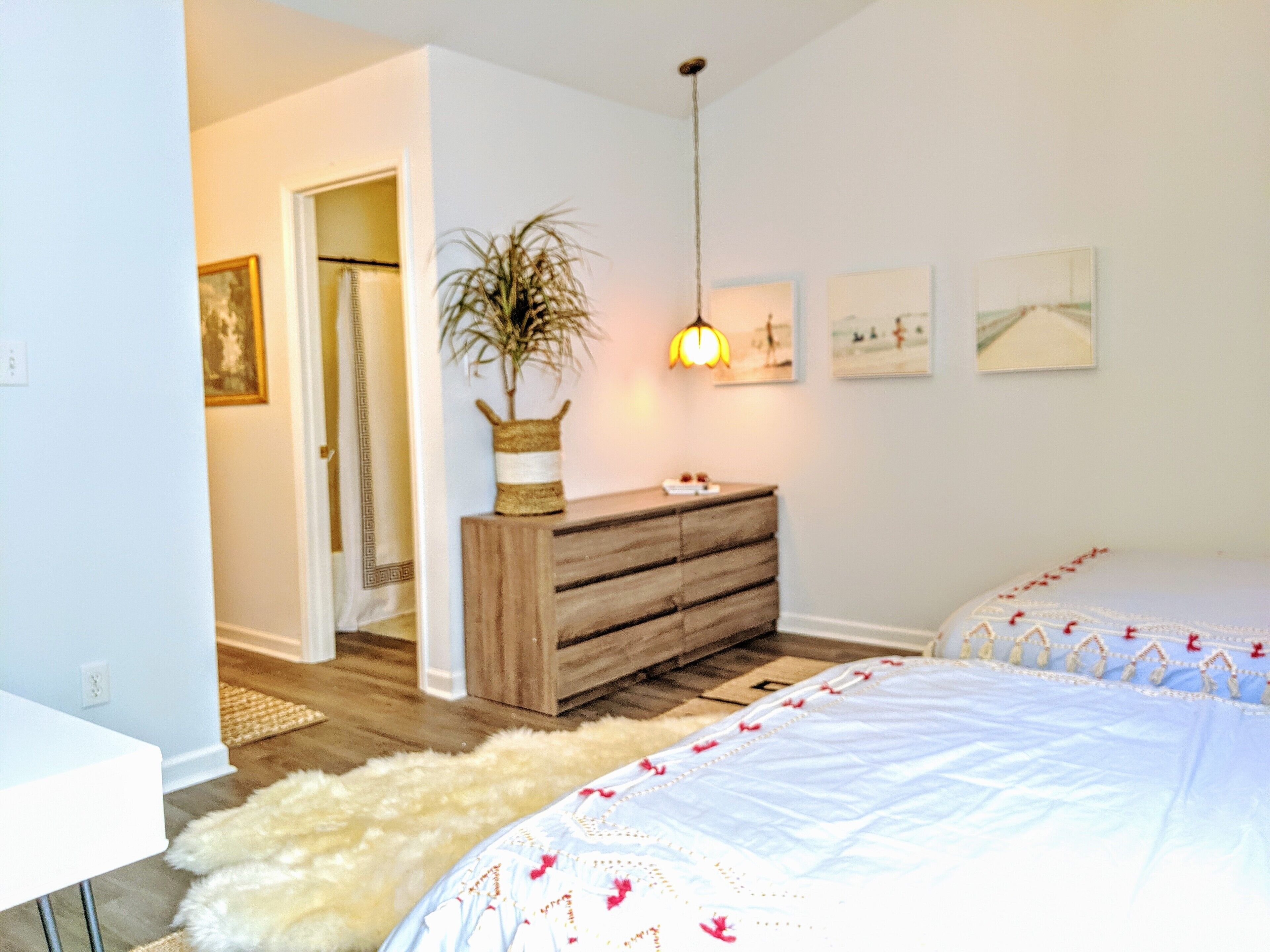 Un dormitorio con un aparador de madera, una cama blanca con ropa de cama floral, una alfombra mullida y una planta colgante.