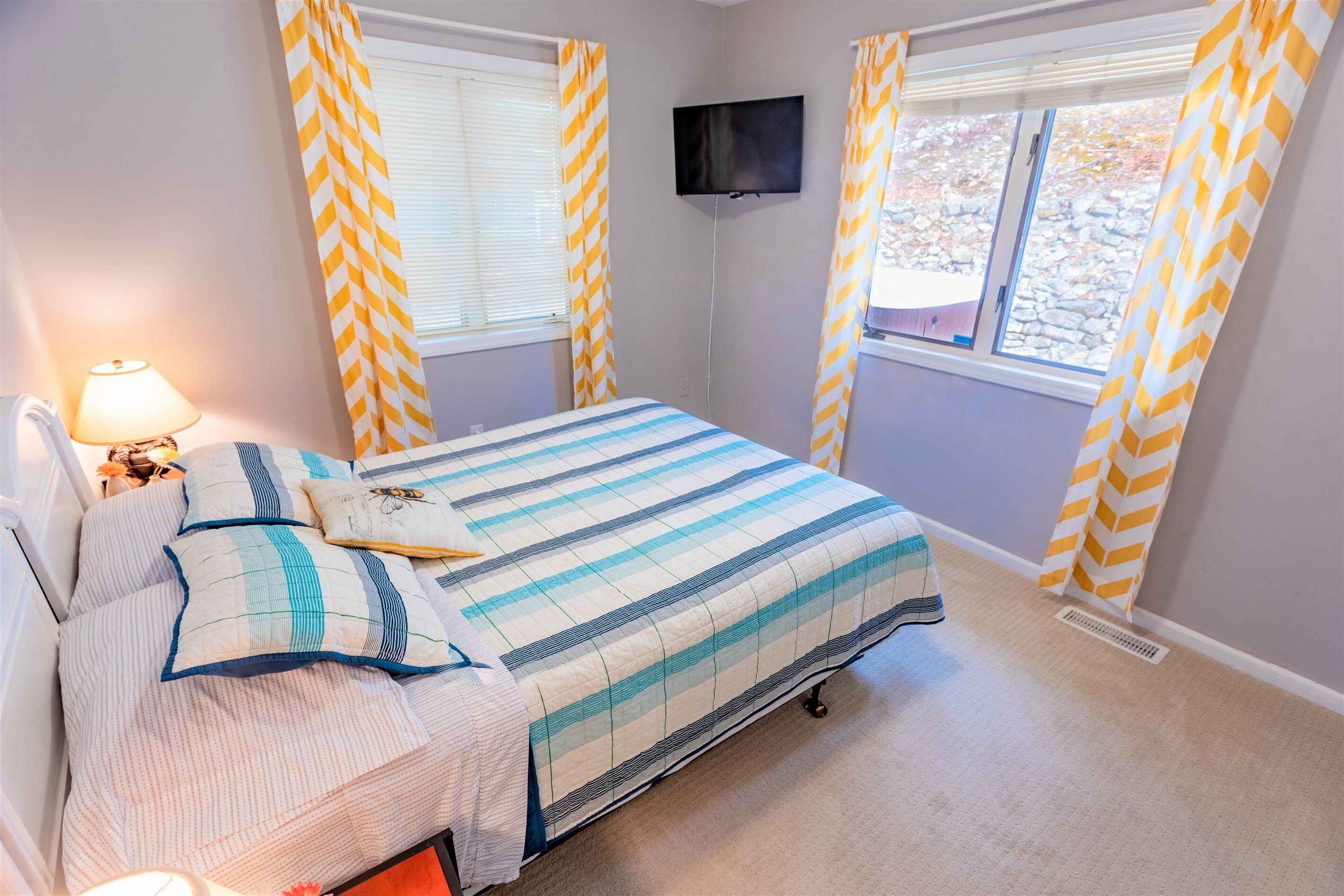 Un dormitorio con cama, televisor montado en la pared y ventanas con cortinas a cuadros amarillos y blancos.