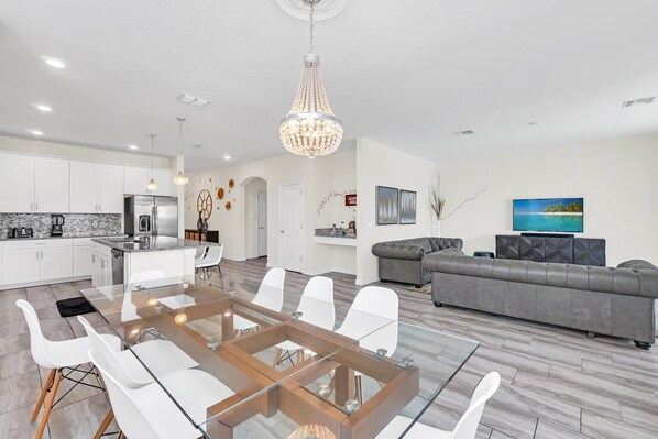 A modern kitchen with a dining area, a living room with a gray sofa, and a large chandelier.