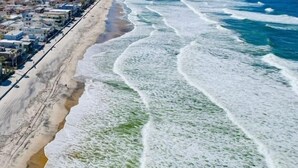 An aerial view of a beach with waves crashing onto the shore, a sandy beach, and buildings along the coastline.