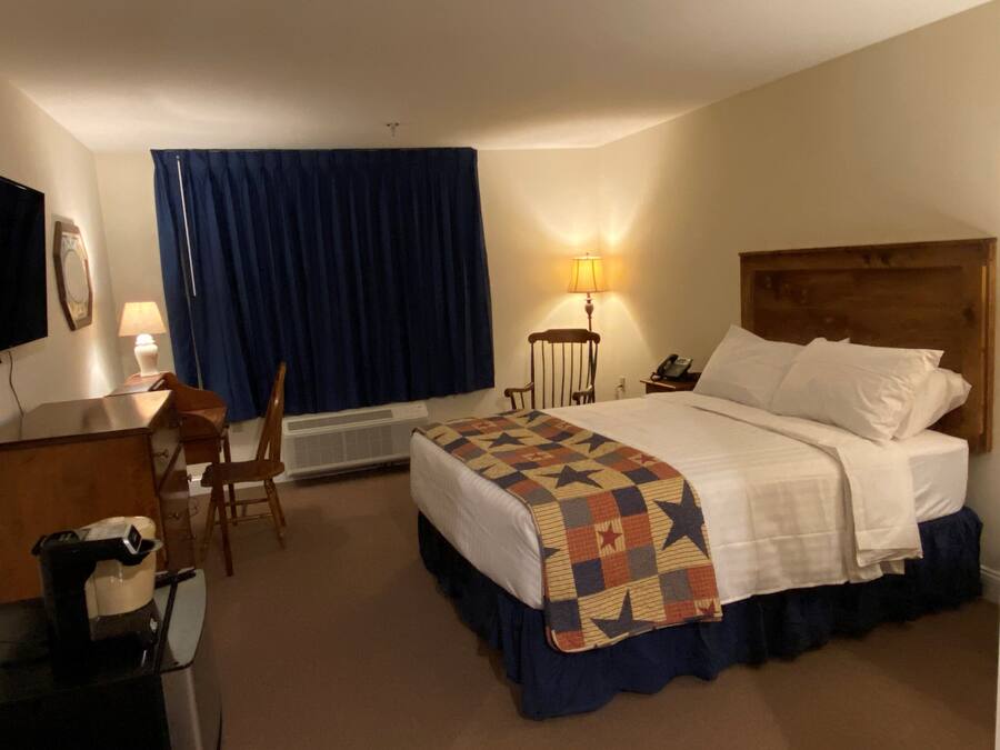 Quarto de hotel com cama, escrivaninha, cadeira, abajur e televisão.