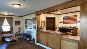 A living room with a wooden bench, a small table with a lamp, a blue chair, and a TV stand with a computer and decorative items. - Country Inn of Hazlet (Hazlet)