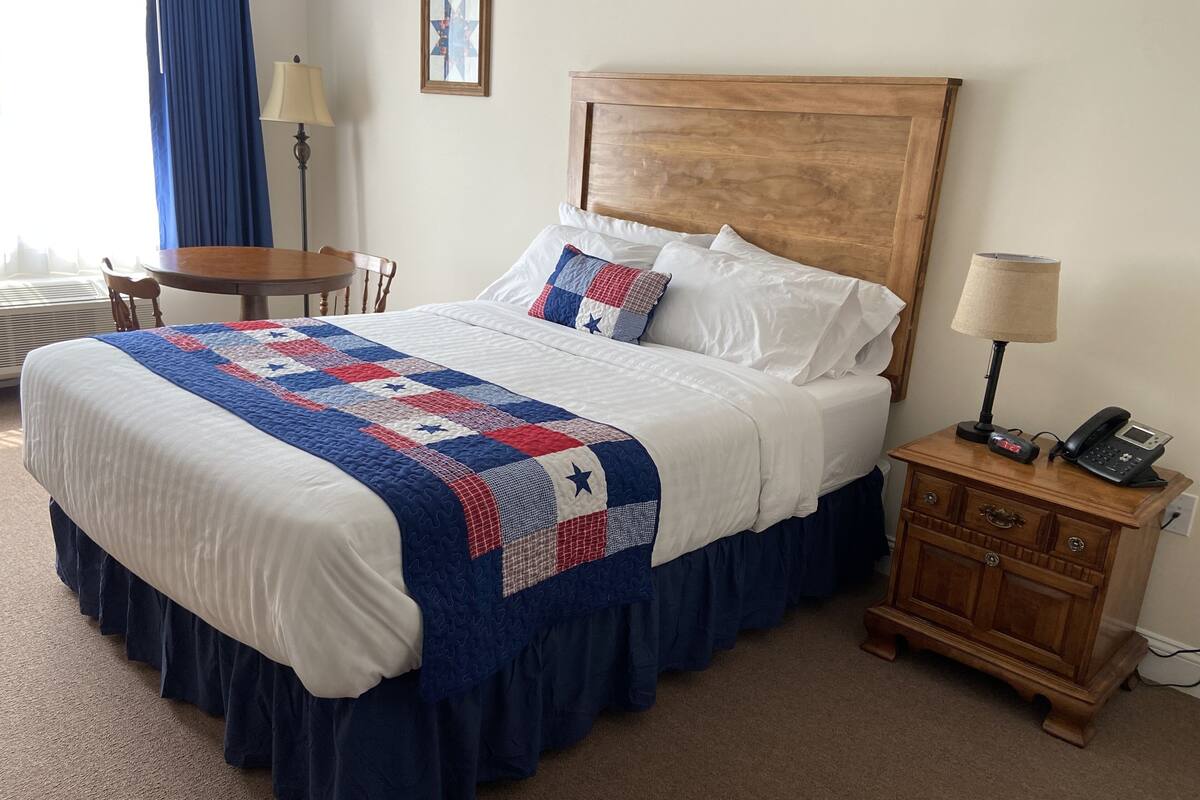 Una cama bien hecha con colcha, una mesita de noche con lámpara y teléfono, y una silla en una habitación de hotel.