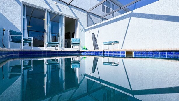 Una piscina con azulejos azules cristalinos, un muro exterior blanco y una puerta que da a un área cubierta con sillas y una mesa.