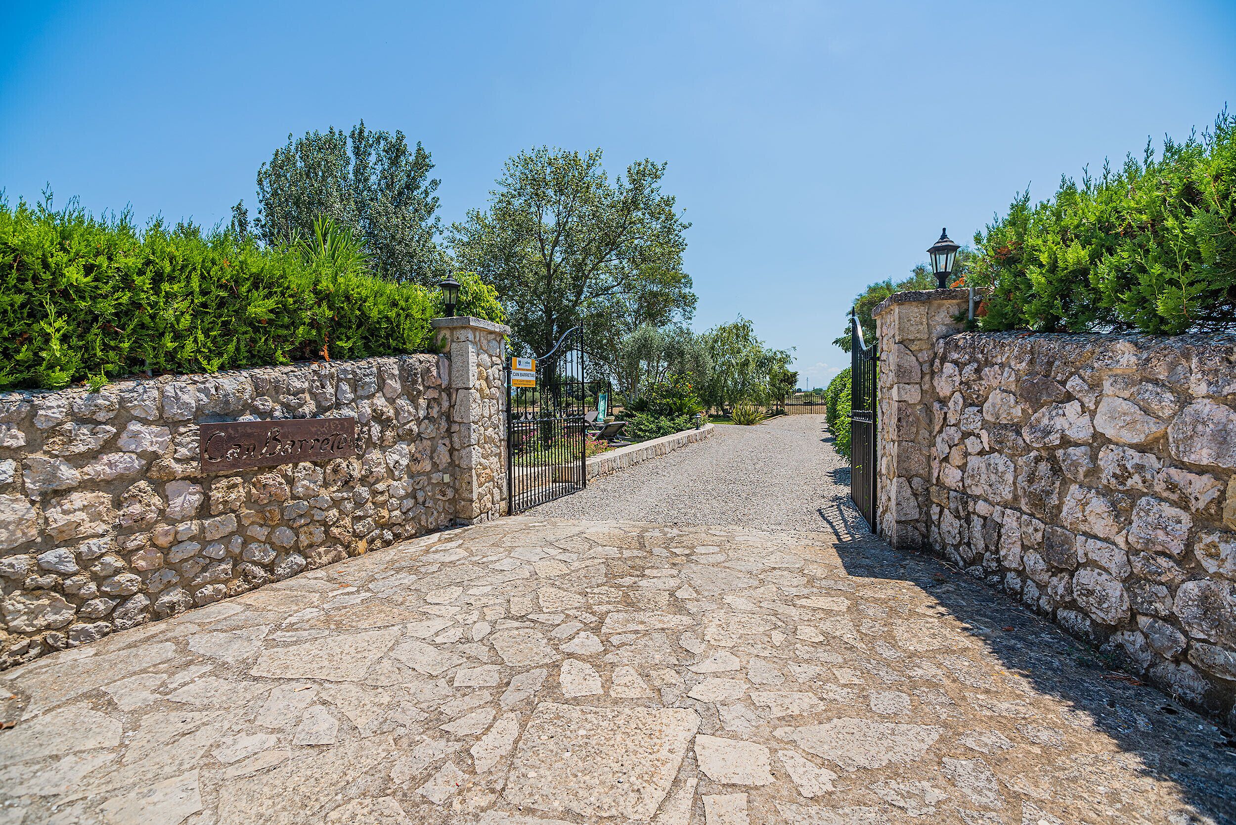 Un chemin de pierres menant à un portail surmonté d’une enseigne, le tout entouré de verdure et d’arbres.