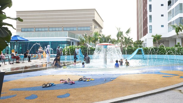 Un área de juegos acuáticos con toboganes y chorros de agua, rodeada de palmeras y un edificio con grandes ventanales.