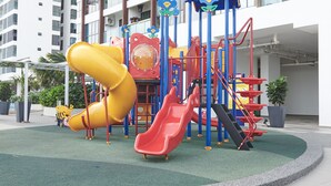 Un parque infantil con toboganes y estructuras para escalar, rodeado por un edificio con balcones y unidades de aire acondicionado.