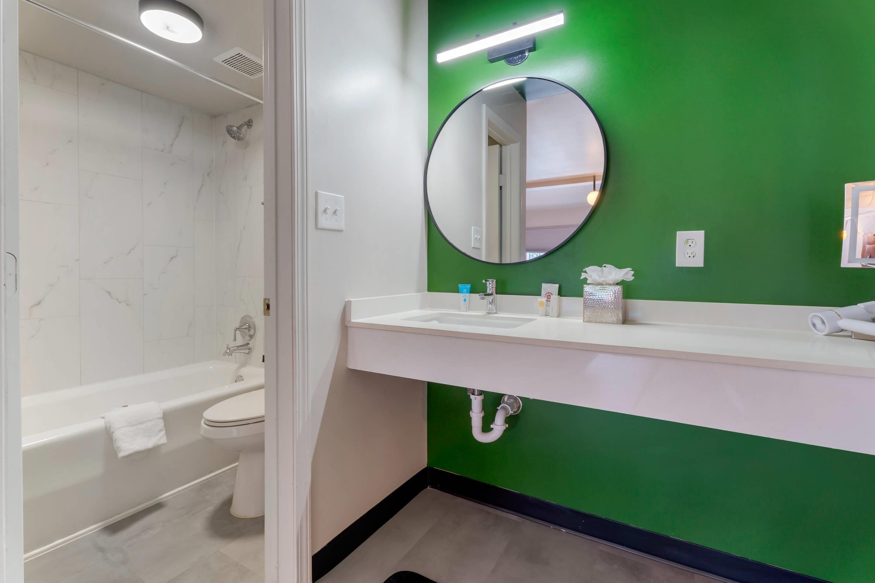 Bagno con una vasca da bagno bianca, un water bianco, una parete verde, uno specchio rotondo e un lavandino bianco con articoli da toilette.