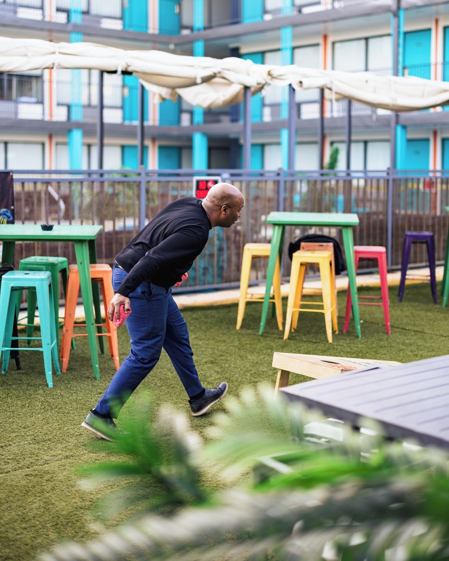 A man walking on a grassy area with colorful chairs and tables, a building with blue and white exterior in the background.
