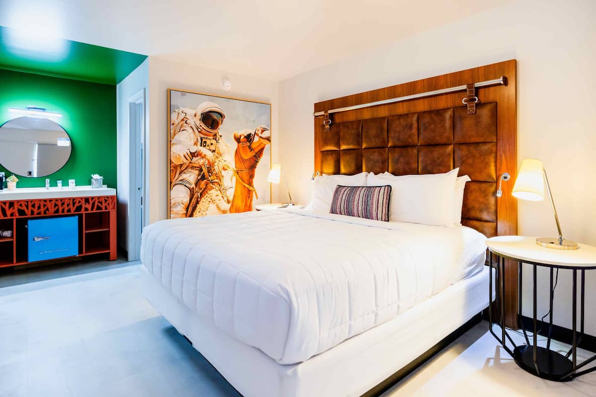 A hotel room with a large bed, a wooden headboard, a round mirror, a painting, a bedside table, and a nightstand.