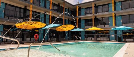 A swimming pool with multiple umbrellas, surrounded by a multi-story building with balconies.