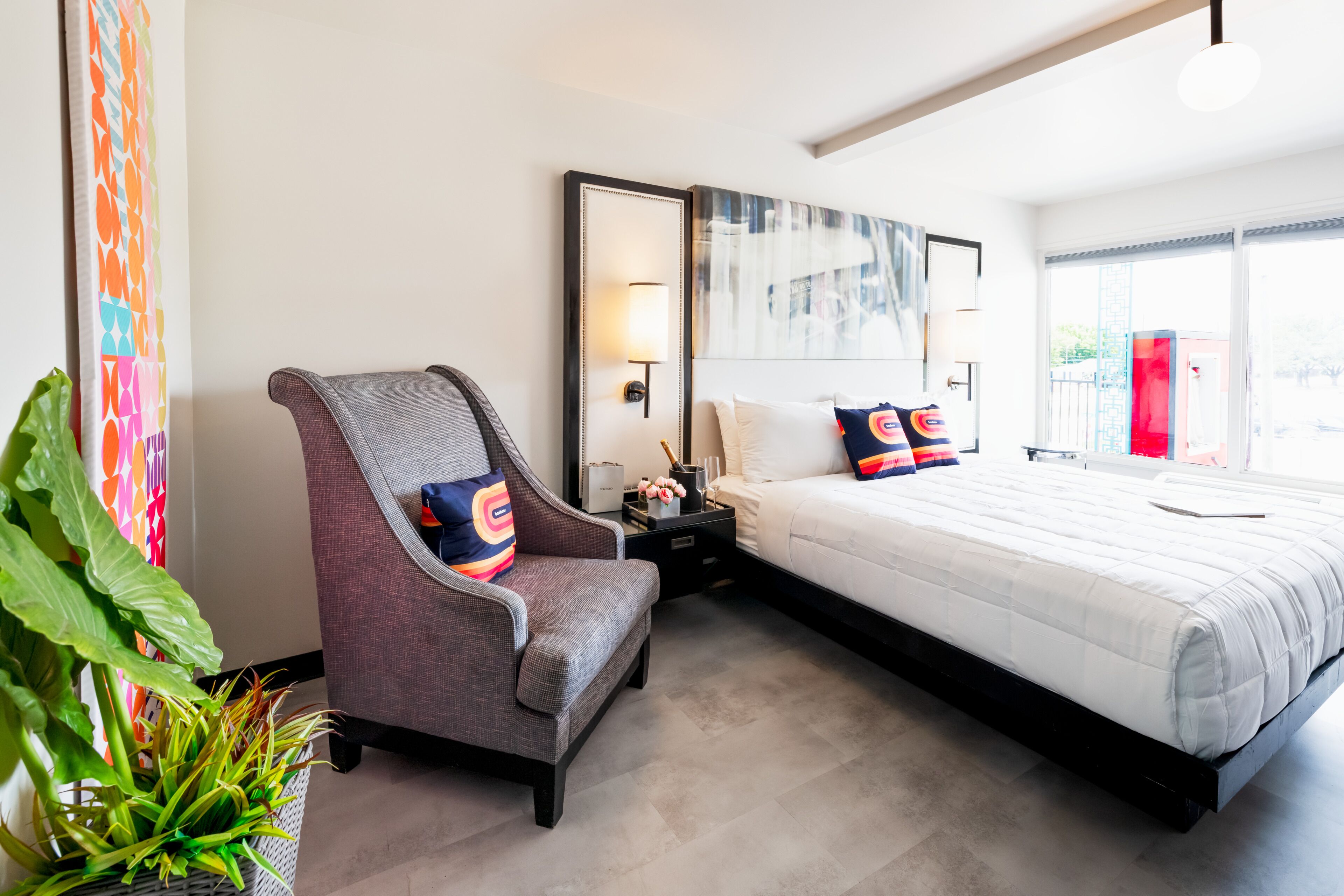 A modern hotel room with a large bed, a grey armchair, a nightstand, a mirror, and a large window with a view of the outside.