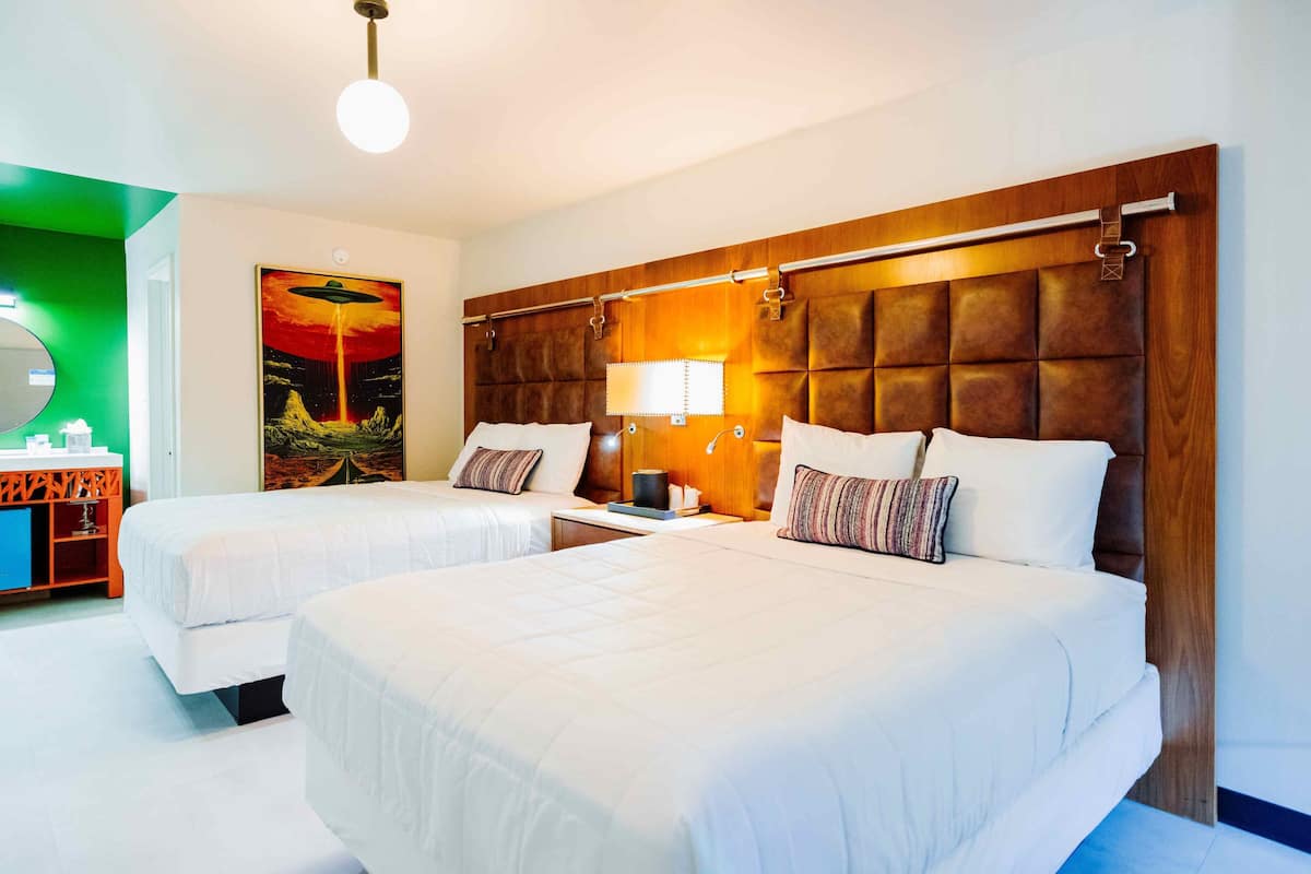 A hotel room with two beds, a wooden headboard, a nightstand, a lamp, and a painting on the wall.