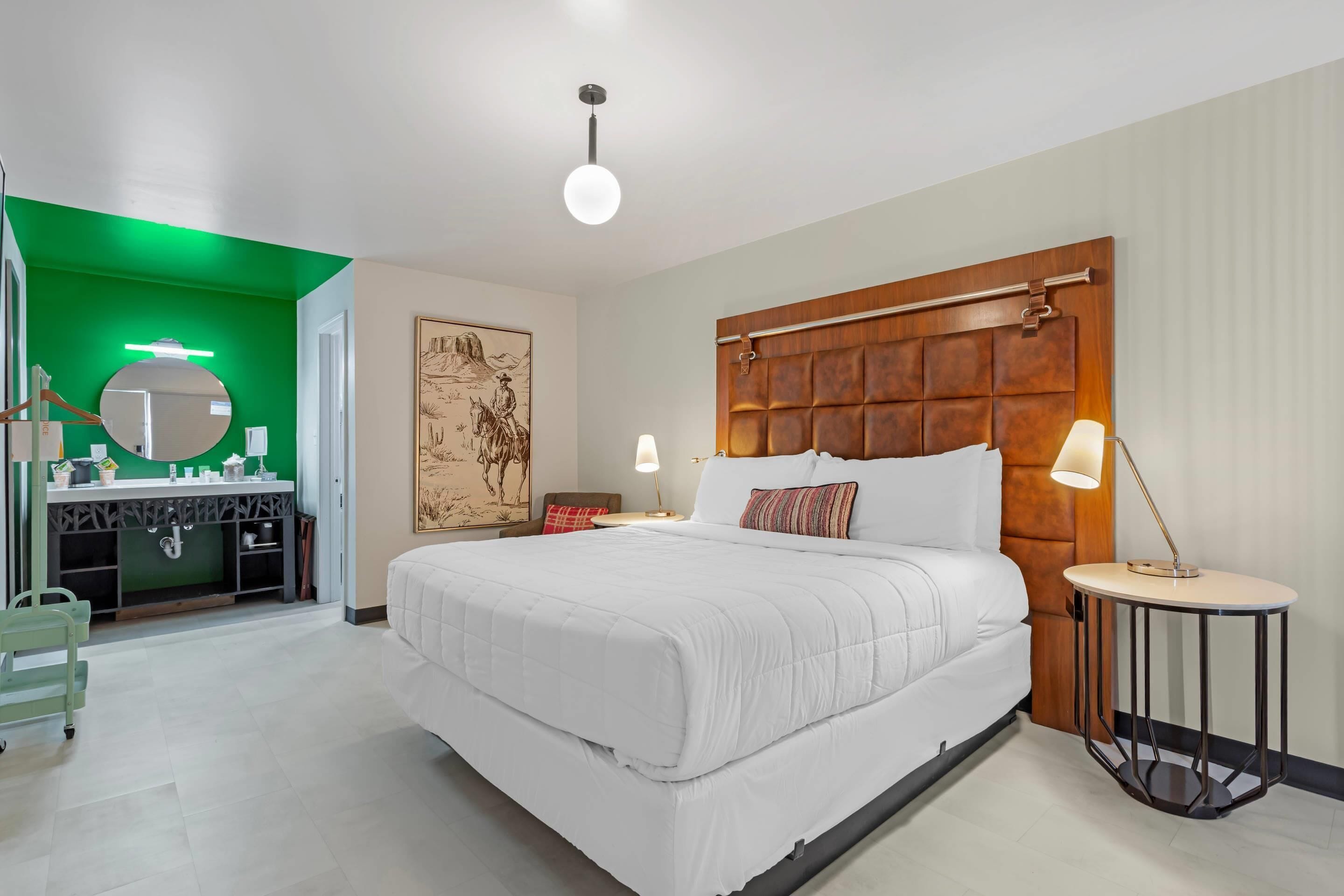 A modern hotel room with a large bed, a wooden headboard, a round mirror, a bedside table with a lamp, and a green accent wall.