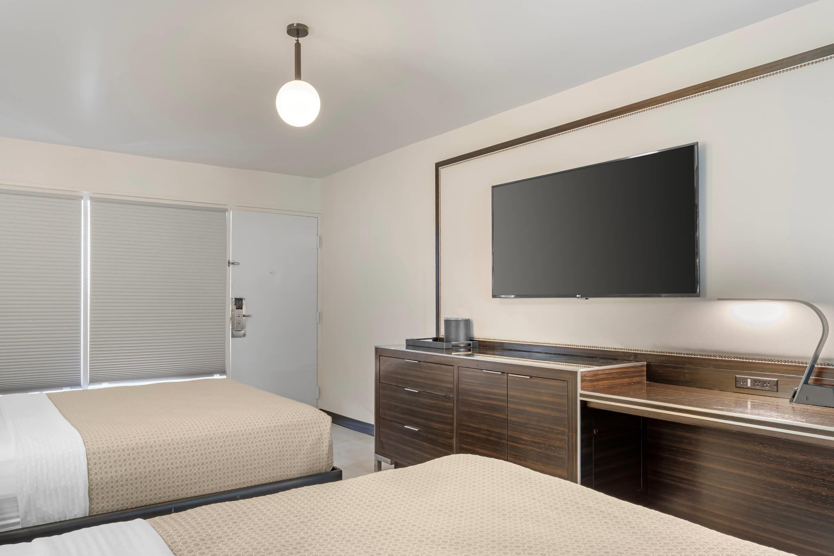 A hotel room with two beds, a desk with a mirror, a flat-screen TV, and a ceiling light.